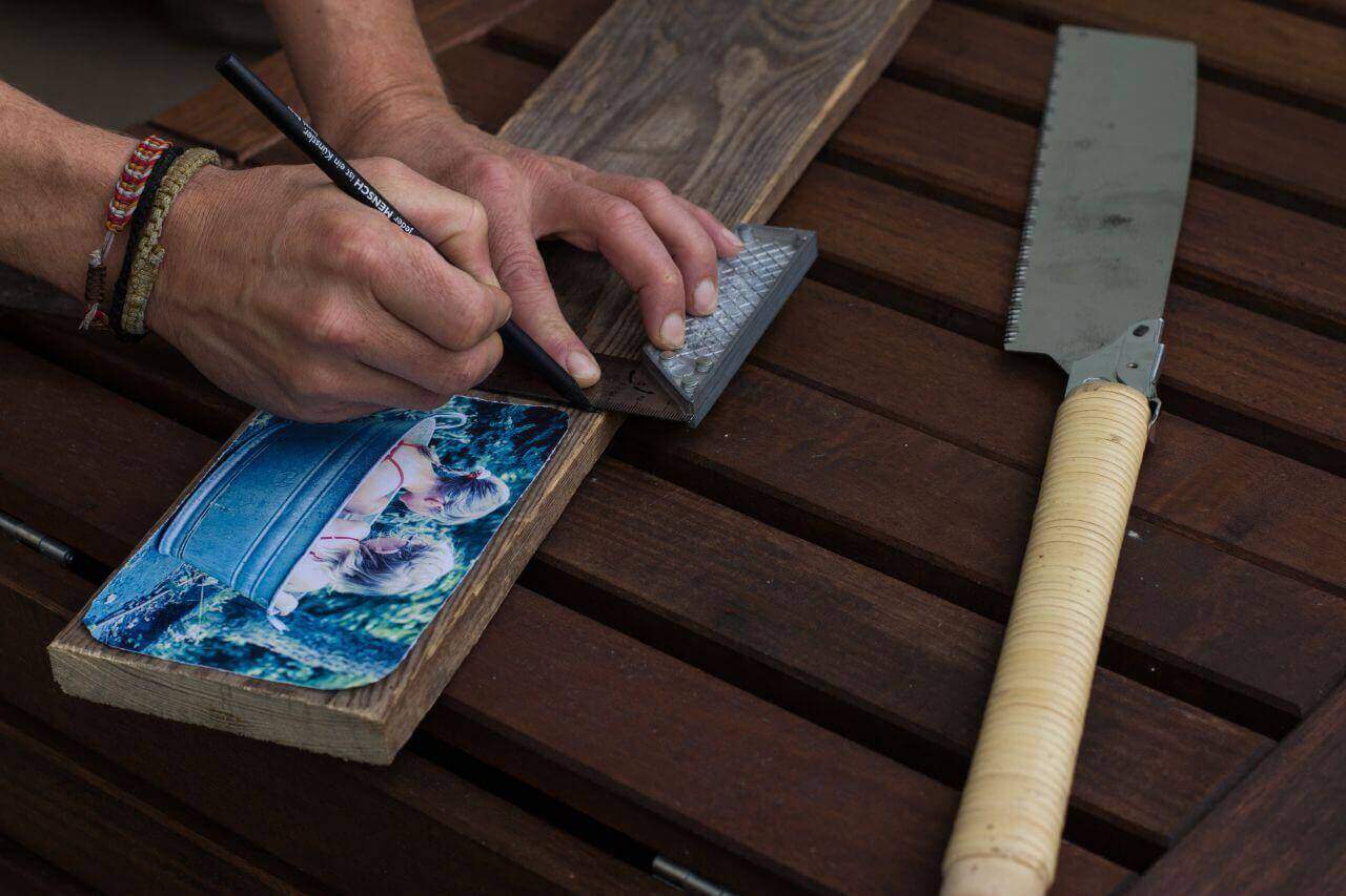 Disegna con un goniometro e una matita le dimensioni della foto sul legno. Sul tavolo si trova anche il seghetto per legno. Disegna con un goniometro e una matita le dimensioni della foto sul legno. Sul tavolo si trova anche il seghetto per legno.
